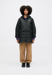 Casaco preto acolchoado com capuz, com grandes bolsos frontais e um logótipo no peito esquerdo; usado sobre um suéter roxo e calças bege.