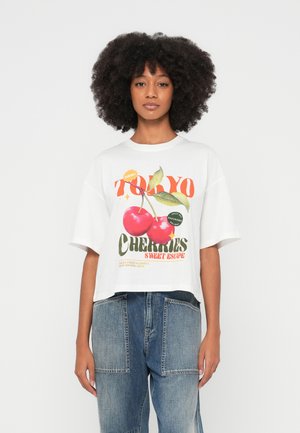 Camiseta blanca corta con estampado gráfico colorido de cerezas y texto, con mangas cortas y cuello redondo. Se lleva con jeans de mezclilla azul.