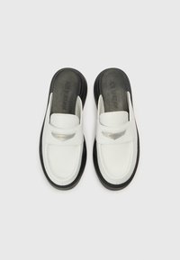 Pantofi slip-on din piele albă cu talpă neagră. Au o curea și un accent argintiu cu inscripția „AIR JORDAN”. Textură netedă, formă rotunjită.