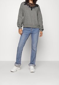 Sudadera gris con detalles de encaje negro en el escote, combinada con jeans azul claro con puños enrollados y zapatillas blancas.