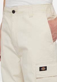 Beige cargobroek gemaakt van duurzaam materiaal met een zichtbare zwarte knop, zijzak en een logopatch met een cirkelvormig ontwerp.