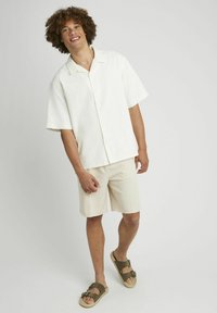 Chemise blanche à manches courtes avec un motif texturé, short beige et sandales à glisser marron avec une boucle, le tout sur fond gris.