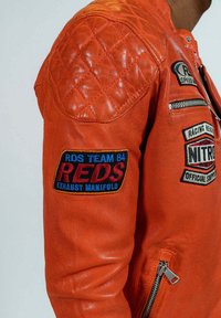 Veste en cuir orange avec des détails matelassés aux épaules, ornée de plusieurs patches colorés et d'une poche zippée sur le bras. Texture lisse.