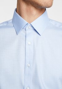 Lichtblauwe button-up shirt met een gestructureerd patroon, puntkraag en kleine witte knopen, met subtiele stikdetails.
