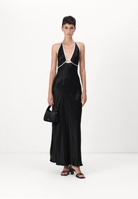 Black satin maxi dress with a plunging neckline, white trim, circular cutout at the chest, paired with a small black handbag and strappy sandals.