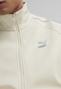 Vit zip-up-jacka med strukturerat tyg, hög krage och ljusblå detaljer med PUMA-logotyp på bröstet.