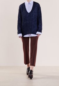Granatowy sweter dziany z dekoltem w kształcie litery V, noszony na białej koszuli w paseczki w granatowym kolorze. Brązowe dopasowane spodnie i czarne buty na sznurówki dopełniają strój.
