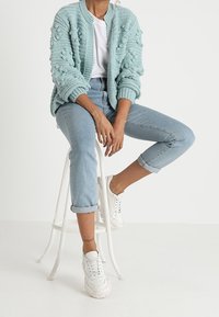 En person sitter på en vit pall, klädd i en ljusblå chunky cardigan, vit t-shirt och uppvikta ljusa jeans i kombination med vita sneakers.