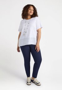 Biała bluzka z lnianym wykończeniem, w zestawieniu z granatowymi, dopasowanymi spodniami i białymi trampkami z czarnymi akcentami, z długim naszyjnikiem z koralików.