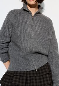 Frau trägt einen grauen, gerippten Reißverschluss-Pullover mit hohem Kragen und einen dunklen, mehrlagigen karierten Rock vor einem schlichten Hintergrund.