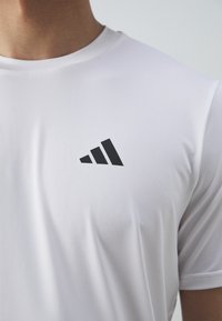 T-shirt de sport blanc avec un col rond et des manches courtes. Présente un logo Adidas noir sur la poitrine. Texture lisse.