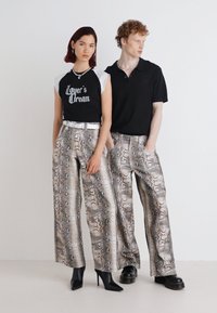 Due modelli stanno fianco a fianco. Uno indossa una camicia bianca e nera con testo stampato e pantaloni a palazzo in stampa serpente; l'altro indossa un polo nera e pantaloni simili.