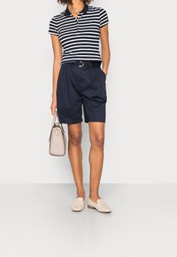 Femme portant un polo à rayures noires et blanches, un short bleu marine avec ceinture, des mocassins beiges, et tenant un sac à main beige, une main dans la poche.