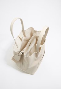 Bolso tote de cuero beige con textura suave, dos asas largas y un compartimento interno con cremallera. Diseño abierto con compartimentos visibles.
