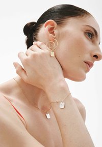 Mujer con el cabello recogido en un moño, usando pendientes espirales de oro, un collar de cadena dorada y una pulsera con colgantes rectangulares, mano tocando el cuello, fondo blanco.