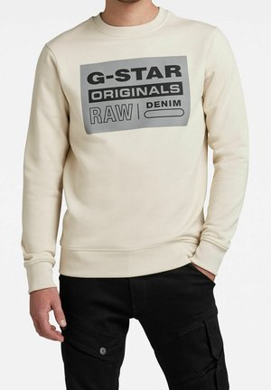 Mężczyzna noszący kremową bluzę z czarno-szarym logo "G-STAR ORIGINALS RAW DENIM" oraz czarne spodnie typu cargo, ręka w kieszeni.