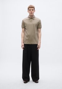 Polo bege com gola, mangas curtas e um logótipo discreto, combinado com calças soltas pretas e sapatos castanho-escuros. Design simples e limpo.