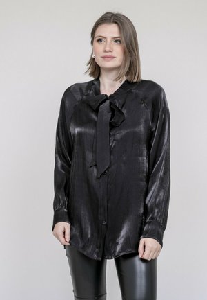 Blouse en satin noir avec un col à nœud, manches longues et fermeture à boutons. Texture lisse avec un éclat subtil, conçue pour un ajustement ample.