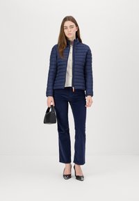 Marineblauwe pufferjack met een gestikt ontwerp, staande kraag en een rits aan de voorkant. Gecombineerd met marineblauwe corduroy broek en zwarte schoenen, terwijl hij een kleine tas vasthoudt.