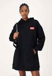 Vestido hoodie preto feito de um tecido suave, com um bolso frontal e um patch do logótipo "DIESEL" em vermelho no peito. Textura suave, corte solto.