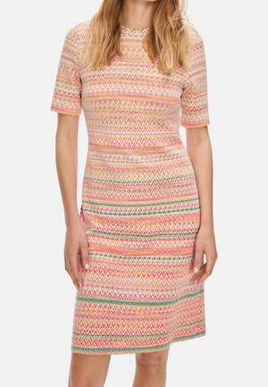 Kobieta w sukience sięgającej kolan z krótkim rękawem, ozdobionej wielokolorowym wzorem zygzaków w odcieniach różu, pomarańczu, zieleni i bieli.