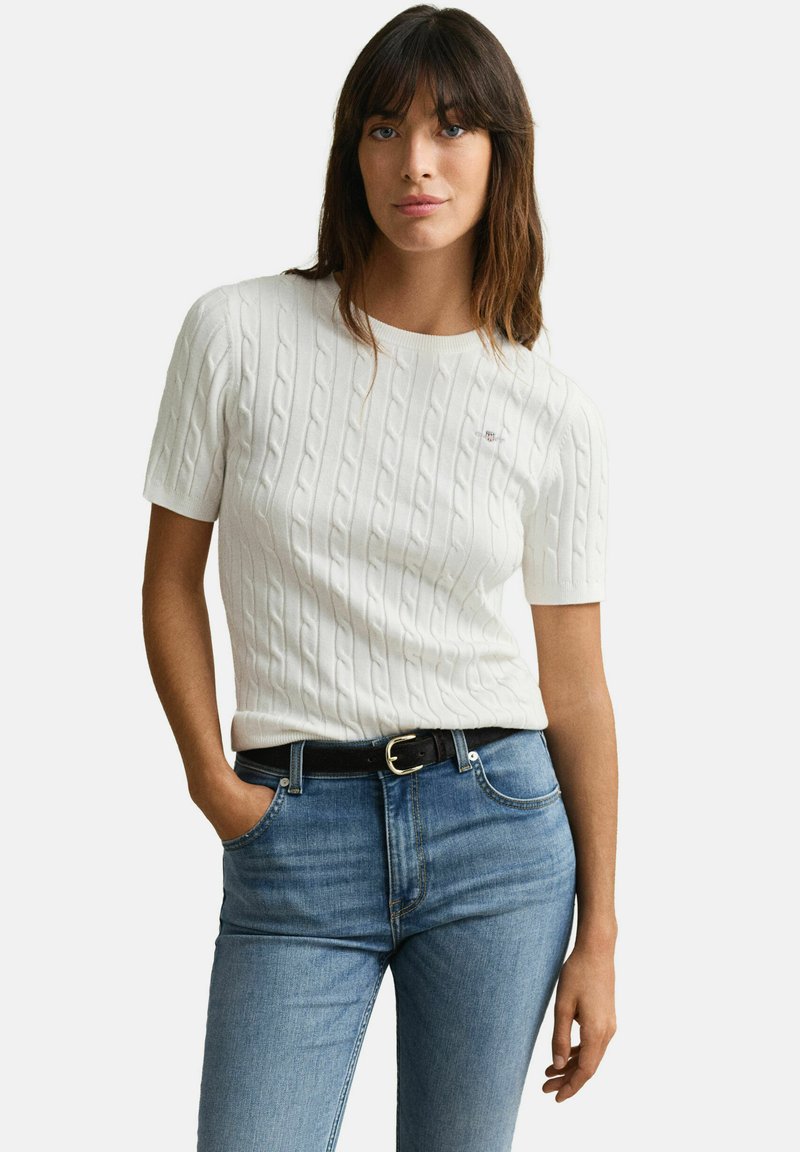 Mujer con suéter blanco de manga corta con tejido de cable y jeans azules con cinturón negro, de pie con una mano en el bolsillo.