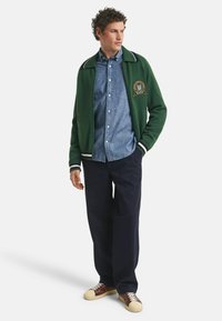 Grøn uldblanding varsity jakke med hvid accentkrave, en blå chambray skjorte med knapper, mørke bukser og multicolor sneakers.