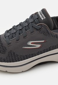 Zapatilla deportiva de color gris oscuro con parte superior de malla, diseño de cordones, suela acolchada y un logo plateado en el lateral. Presenta acentos texturizados.