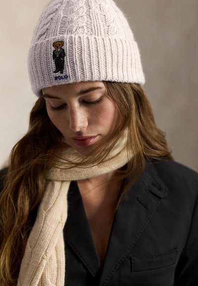 Világos rózsaszín kötött sapka fonott mintával, macis öltönyös folttal és "POLO" márkajelzéssel; krémszínű sállal párosítva.