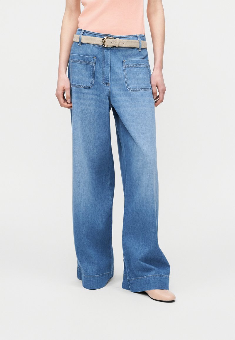 Jean en denim bleu à jambes larges avec poches plaquées devant, ceinture beige, porté par une personne en haut rose clair et chaussures beiges sur fond blanc.