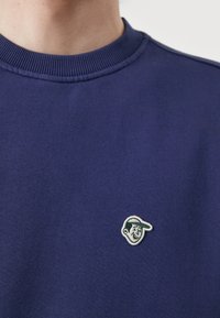 Felpa blu navy con colletto a coste e un piccolo logo a faccia ricamato in bianco e verde sul petto. Tessuto morbido e dalla texture piacevole.