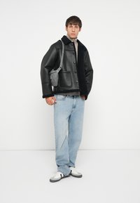 Schwarze Lederjacke mit Schafslederkragen, graue Umhängetasche, hellblaue Jeans und weiße Sneakers. Das Model steht vor einem schlichten Hintergrund.