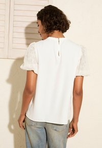 Top bianco con maniche corte in pizzo floreale, dettaglio a forma di chiave sulla schiena e una finitura liscia. Abbinato a pantaloni in denim azzurro chiaro.
