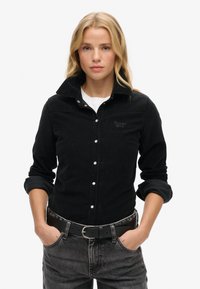 Sort fløjlsskjorte med knaplukning fortil, krave og diskret logo; parret med et sort bælte og grå denim jeans.