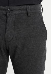 Black textured trousers with a button closure, featuring a pleated front and side pockets, made from a lightweight fabric.