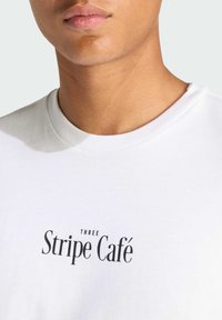 Biała bawełniana koszulka z czarnym nadrukiem logo, na którym widnieje napis "THREE Stripe Café." Okrągły dekolt o gładkiej fakturze.