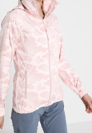 Vrouw met een lichtroze camouflageprint ritsjack met hoge kraag en blauwe broek tegen een effen achtergrond.