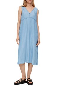 Jasnoniebieska dżinsowa sukienka bez rękawów z dekoltem w kształcie litery V, długości do kolan, z bocznymi rozcięciami i subtelnymi detalami w szyciu. Zestawiona z czarnymi sandałami na paski.