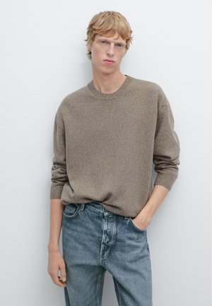 Hellbrauner Strickpullover mit Rundhalsausschnitt, getragen zu hellblauen Jeans. Der Pullover hat aufgerollte Ärmel und einen lockeren Schnitt.
