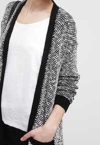 Vzorovaná pletená cardigan v černobílé barvě s texturovaným designem, dlouhými rukávy a černým lemem. Stylizováno s jednoduchým bílým tričkem pod ním.