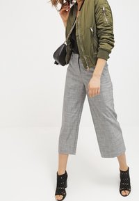 Oliverově zelená bomber bunda, černý top, šedé kostkované kalhoty do poloviny lýtek a černé polobotky s otevřenou špičkou a výřezy.