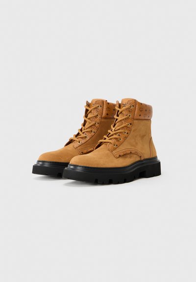 Tan suede ankle boots with a black rubber sole, featuring lace-up design and decorative hardware on the collar. Textured accents on upper.