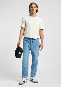 T-shirt crema con taschino, pantaloni a righe blu, cappellino nero in mano, sneaker bianche con accenti neri e grigi, vestibilità rilassata e look informale.