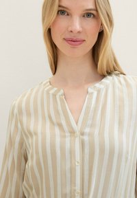 Blouse rayée beige et blanche avec un col, fermeture à boutons et tissu léger. Présente des rayures verticales et une coupe décontractée.