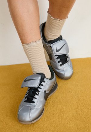 Personne portant des chaussettes côtelées beiges et des baskets Nike argentées avec des lacets noirs et une bride ajustable sur un tapis moutarde.