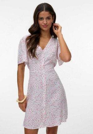 Kobieta z długimi falującymi brązowymi włosami, ubrana w białą sukienkę z krótkim rękawem z różowym kwiatowym wzorem i beżową bransoletką na lewym nadgarstku.