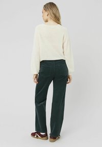 Un pull crème clair à texture côtelée, associé à un pantalon large en velours côtelé vert foncé. Des baskets bordeaux avec des accents blancs complètent la tenue.