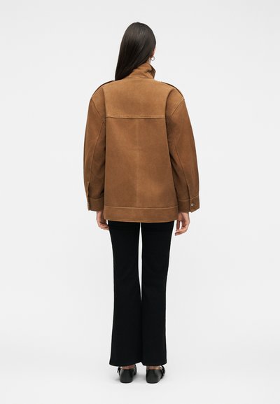 Γυναίκα με μακριά σκούρα μαλλιά, φορώντας καφέ μπουφάν, μαύρο φαρδύ παντελόνι και μαύρα παπούτσια, στέκεται πλάτη σε φόντο χωρίς σχέδια.