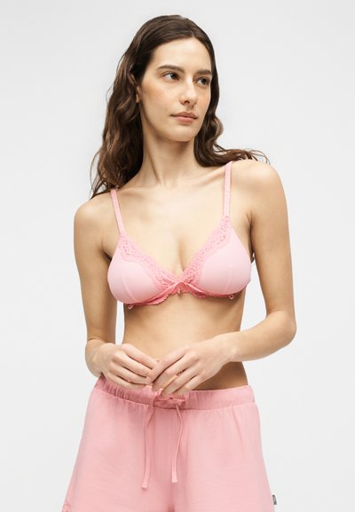 Γυναίκα με μακριά καστανά μαλλιά που φοράει ένα ροζ σουτιέν με δαντέλα και ροζ σορτς με κορδόνι σε απλό φόντο.