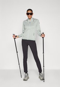 Veste zippée à capuche vert clair à motif, pantalons noirs et chaussures de randonnée grises ; tient des bâtons de trekking et porte des lunettes de soleil noires.